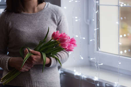 Young woman holding tender bouquet of pink tulips near window with magic bokeh lights. Spring gift, with copy space. Mother day present.の写真素材