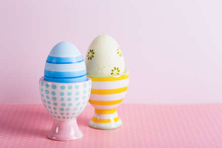 Closeup two colorful painted Easter eggs in vibrant modern egg stands on pastel pink background. Spring home minimalistic composition with copy space.の写真素材