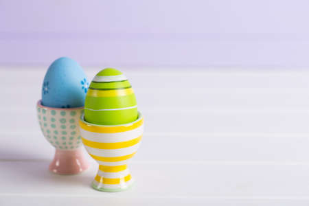 Closeup two colorful painted Easter eggs in vibrant modern egg stands on pastel lilac background. Spring home minimalistic composition with copy space.の写真素材
