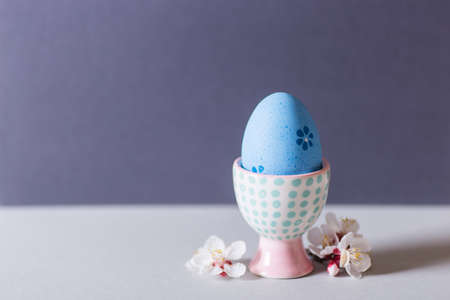 Closeup colorful painted Easter egg in vibrant modern egg stand surrounded by white flowering tree branches on gray background. Home minimalistic composition with copy space.の写真素材