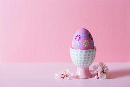 Closeup colorful painted Easter egg in vibrant modern egg stand surrounded by white flowering tree branches on pastel pink background. Home minimalistic composition with copy space.の写真素材