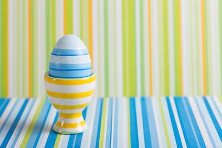 Closeup colorful painted Easter egg in vibrant modern egg stand on striped green and blue background. Spring home minimalistic composition with copy space. Geometric style.の写真素材
