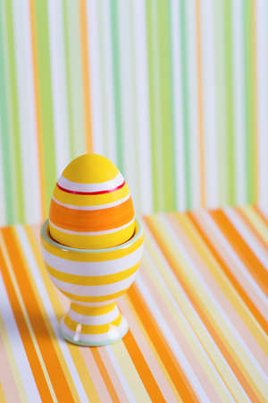 Closeup colorful painted Easter egg in vibrant modern egg stand on striped green and orange background. Spring home minimalistic composition with copy space. Geometric style.の写真素材