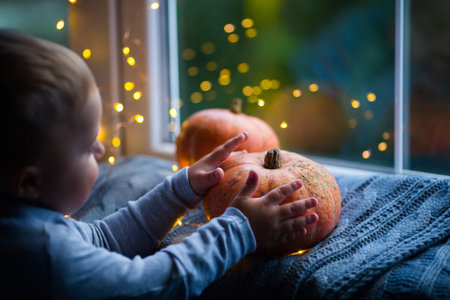Toddler boy holding orange pumpkin on gray knitted plaid near window in evening surrounded with warm garland lights with golden bokeh. October autumn harvest, home decoration for Halloween.の写真素材