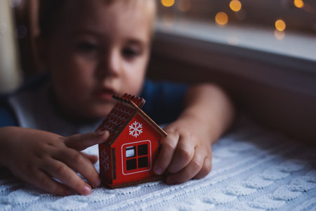 Little child boy holding red decorative wooden Christmas house on white knitted background. Cute...