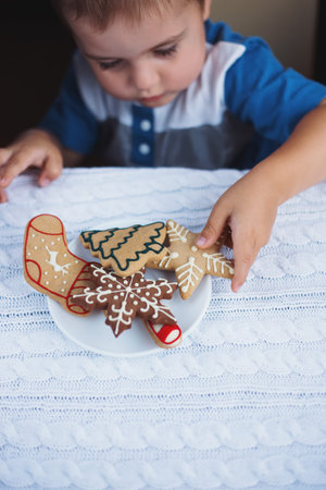 Child boy taking Christmas gingerbread from plate on table near window in daylight. New Year glazed...
