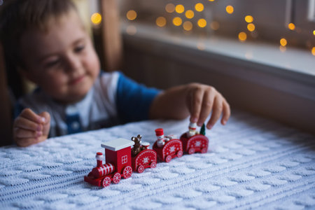 Child boy playing with red toy Christmas train on white knitted plaid in daylight with warm garland bokeh background. Cozy home New Year atmosphere.の写真素材
