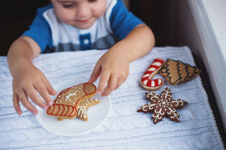 Smiling child boy taking Christmas gingerbread from plate on table near window in daylight. New Year glazed decorated cookie. Cozy home atmosphere.の写真素材