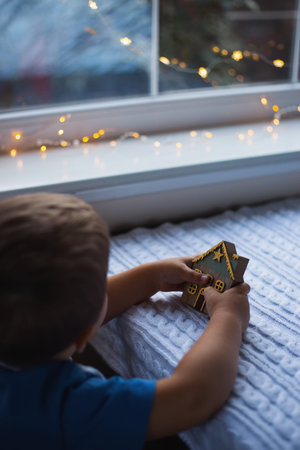 Child boy holding decorative wooden Christmas house on white knitted background. Cute festive home decor. Cozy New Year atmosphere.の写真素材