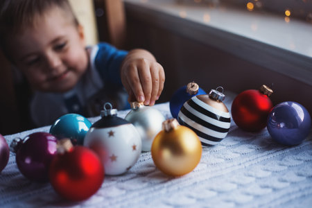 Happy child boy choosing Christmas ball near window indoor with warm garland lights on blurred...