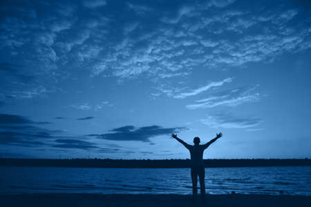 Silhouette of man with arms outstretched against cloudy sky on evening near lake. Natural banner with color of the year 2020 - Classic Blue. Concept of freedom. Copy space.の写真素材