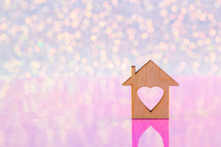 Wooden icon of house with hole in form of heart on glitter shiny light and pink background with bokeh lights. Festive romantic banner with copy space. Concept of sweet homeの写真素材