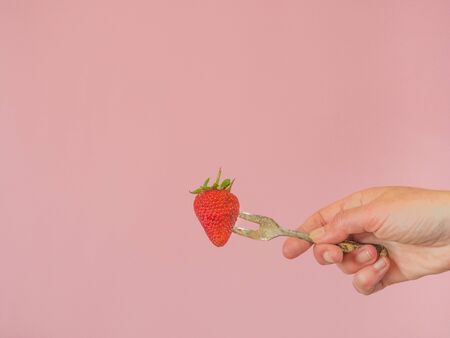 Hand with strawberry and old fork on pink background with copy spaceの写真素材