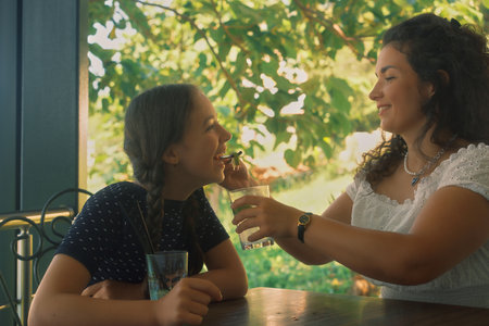 A mother feeds her daughter a slice of lime from a drink. Green trees in the backgroundの写真素材