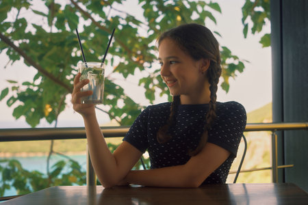 A young girl sitting at a table in a summer cafÃ©, holding a glass of drink in her right hand and looking at itの写真素材