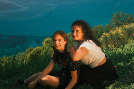 Mother and daughter sit on the bank of a high cliff and look at the camera lens. The sea in the backgroundの写真素材
