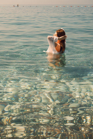 A young girl with her eyes closed, wearing wet clothes, standing chest-deep in water and fixing her wet hairの写真素材