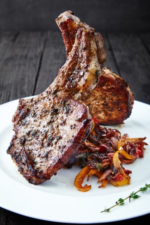 Delicious, fleshy, hot meat ribs with sauteed bell peppers and onions, decorated with a thyme sprig on white plate. Appetizing main dish on black wooden background. Club Steak veal on the bone.の写真素材