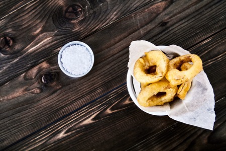 Tasty, crunchy, golden onion rings in a white bowl with a paper. Dark wood background, a cup of salt next to it.の写真素材