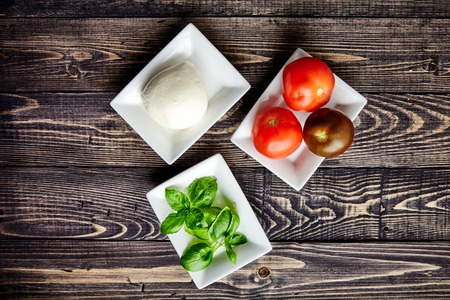 Top view of the main ingredients of Italian Caprese salad, mozzarella cheese, juicy tomatoes and fresh Basil in white plates. Traditional Mediterranean cuisine. Popular Italian dish on wooden tableの写真素材