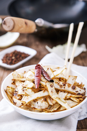 Stir fried cabbage in wok with Sichuan pepper, dried chili and soy sauce in white bowl, decorated with whole pods of chili peppers. Chopsticks and frying pan wok next to it, on wooden tableの写真素材