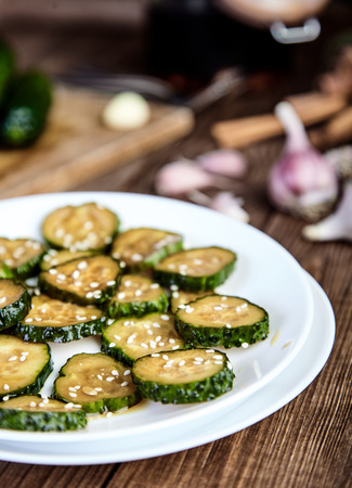 Slices of crunchy pickled cucumbers in soy sauce and rice vinegar on white plate, sprinkled with sesame seeds. Garlic, heap of pickles and cutting board behind. Chinese recipe. Shallow depth of fieldの写真素材