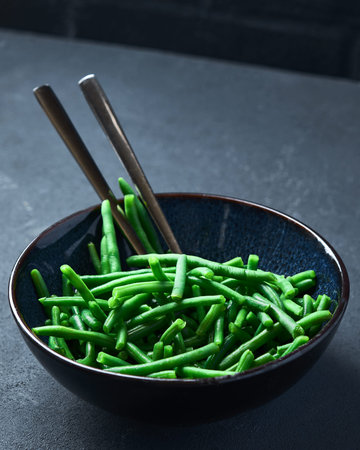Green beans in a black bowl on a gray table. 45 view. There is a spoon in a bowl. Close up