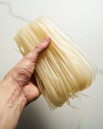 Mans hand holding a wide rice noodles on white and clean marble background. Raw and uncooked, dry.の写真素材