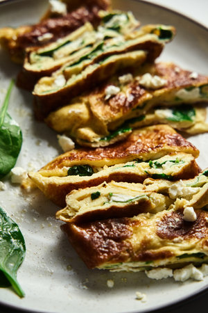 Close up of sliced folded omelette stuffed with spinach and feta on the white plate with spinach leaves on back. Healthy and dietary breakfast dish. Minimalistic food. White marble background.の写真素材