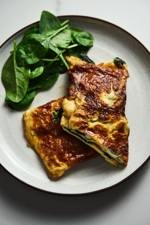 Folded omelette stuffed with spinach and feta on the white plate with spinach leaves on back. Healthy and dietary breakfast dish. Minimalistic food photo. White marble background.の写真素材