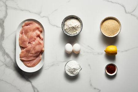 Raw Chicken fillet cut into schnitzel on an oval platter on a white marble kitchen countertop. Preparing for cooking baked chicken schnitzel. Other ingredients nearの写真素材