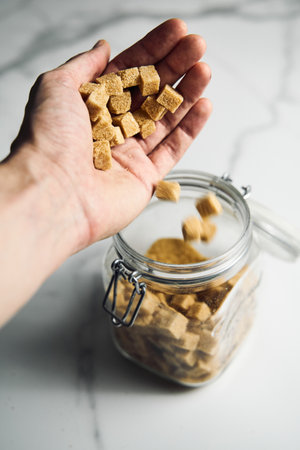 Brown sugar cubes in a mans hand and in the jar on white marble background.の写真素材