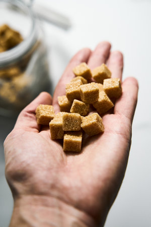 Brown sugar cubes in a mans hand and in the jar on white marble background.の写真素材