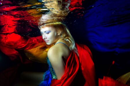 Beautiful girl swims and poses underwater in a blue dress with red, orange cloth over blue background and looking over your shoulder. Close-up portrait. Horizontal orientationの写真素材