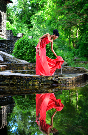 African American girl in a red dress with red shoes in hand is worth in the summer on the water on the stones in the Park on a background of green plants and is reflected in the water.の写真素材