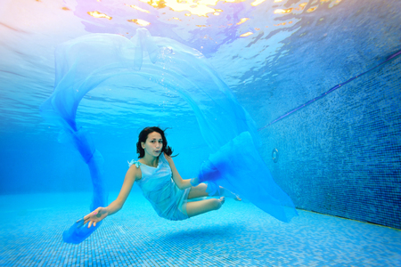 Teen girl swims underwater in the pool on a blue background, looks at the camera and plays with a blue cloth. Portrait. Shooting under water. Landscape orientationの写真素材