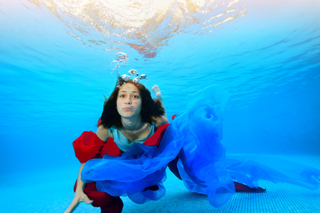 A teenage girl with a red and blue cloth swims underwater near the bottom and looks at the camera. Portrait. Shooting under water. Horizontal orientationの写真素材