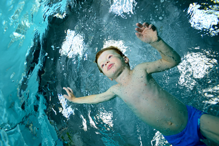 A little boy is engaged in sports swimming in the pool. Swims under water on a blue background and looks forward. Portrait. Shooting under water on a background of superficiality. Horizontal view.の写真素材