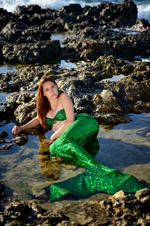 A beautiful teenage girl at sunset in a green mermaid costume lies on the beach among the rocks and looks at the camera. Portrait. Vertical orientation.の写真素材