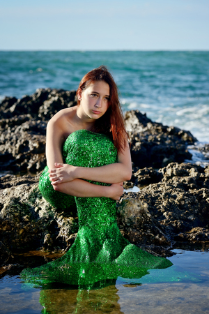 A teenage girl with red hair in a mermaid costume sits on the stones on the beach, hugging her knees and looking to the side. Portrait. Vertical view.の写真素材