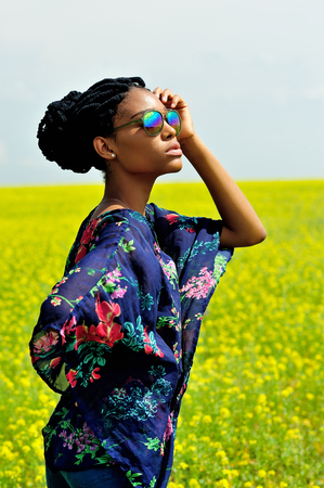 Beautiful African American girl model with dreadlocks is in the field against the background of yellow flowers in sunglasses. She raised her hand to her head and looks at the sun. Portrait.の写真素材