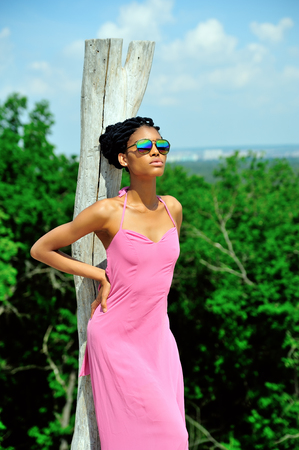 An African-American girl stands near a broken dry tree in a pink dress, with dreadlocks on her head, sunglasses and looking at the sun. Portrait.の写真素材