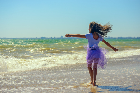 Happy little girl is spinning and dancing on the beach on a Sunny day in a purple dress against the background of raging waves. Her hair is fluttering in the wind. The horizontal orientation.の写真素材