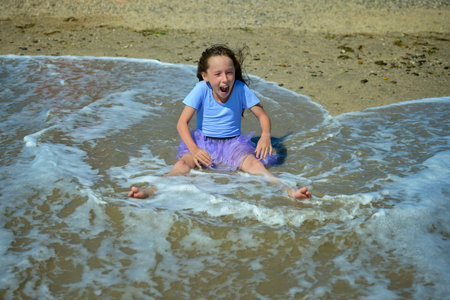 Cheerful little girl sitting on the yellow sand on the beach on a Sunny day in a purple dress, playing and laughing when the wave rolls on her. Horizontal orientation.の写真素材