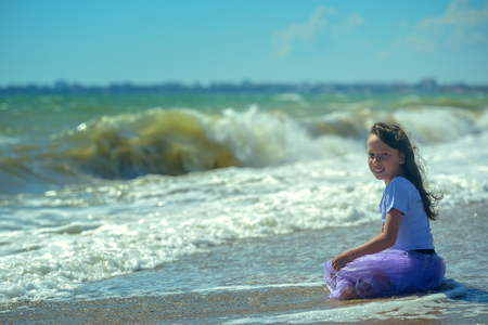 A little beautiful girl in a purple dress sits on her knees at the edge of the water on the beach on a Sunny day, looks at the camera and smiles against the background of raging waves.の写真素材