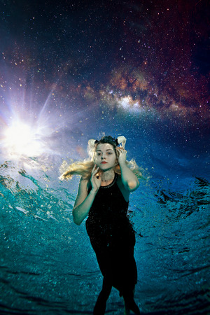 Young beautiful girl with white horns on her head swims in a black dress at night underwater in the pool against the background of space, the night sky and stars. She's looking at the camera. Portraitの写真素材