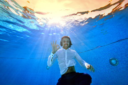 A young smiling man with a headset on his head swims and poses underwater in the pool in a white shirt against the yellow sunset. He's looking at the camera. Portrait. Underwater photography.の写真素材