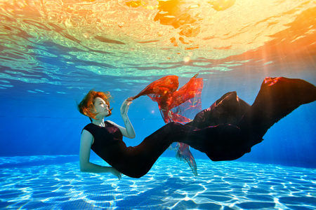 Fantastic young girl with red hair posing underwater in the pool. It sinks to the bottom and plays with a red cloth against the background of a yellow sunset. Underwater photography.の写真素材