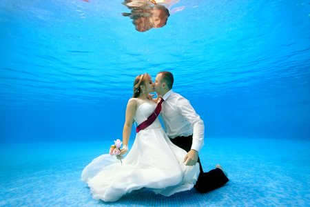 The bride and groom in wedding dresses embrace and kiss underwater at the bottom of a swimming pool. Portrait. Landscape orientation. Shooting under waterのeditorial素材