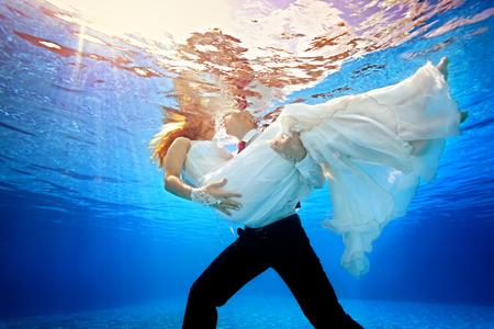 The groom is holding his bride in white dress on background of sun rays underwater in the pool. Horizontal orientation. A view from under the water from the bottom of the poolの写真素材